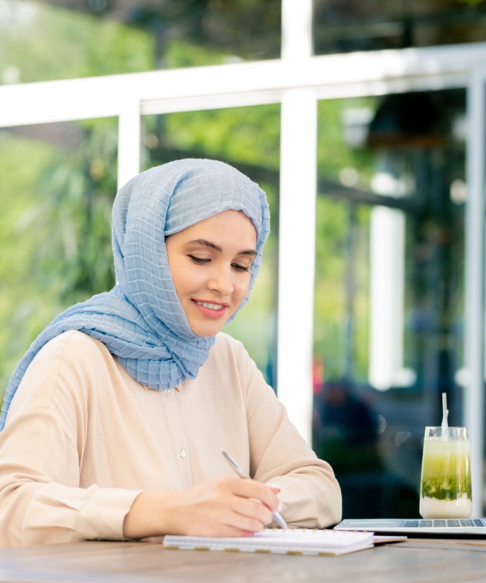 young-muslim-student-in-hijab-making-working-notes-by-table.jpg